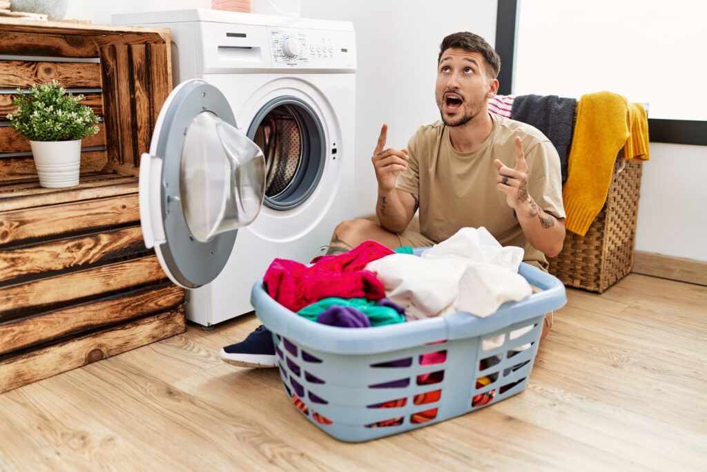 man putting dirty laundry into washing machine amazed and surprised looking up and pointing with fingers and raised arms.